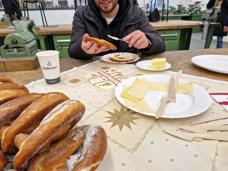 Ein Mann sitzt an einem Tisch und bestreicht eine Brezel mit Butter. Im Vordergrund sind mehrere Brezeln zu sehen und einen Teller mit Käsescheiben und Holzgabeln. Auf dem Tisch stehen außerdem ein Pappbecher und ein Teller mit Butter. Die Szene scheint sich in einem Werkraum abzuspielen, denn im Hintergrund befinden sich Werkbänke mit Schraubstöcken.